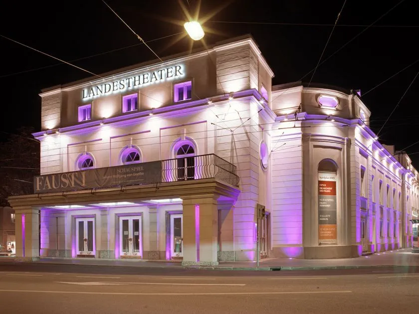 Night view of the Landestheater Salzburg facade illuminated with purple lighting, showing the LANDESTHEATER sign and a FAUST banner