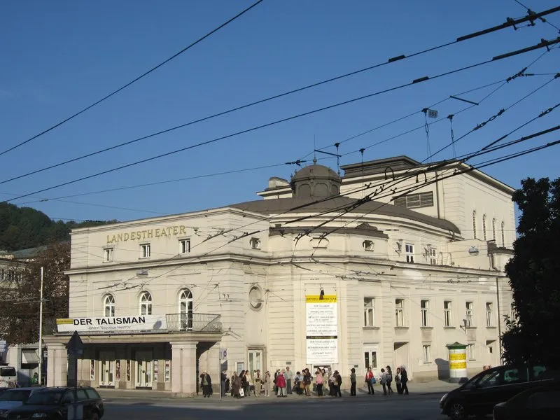 Exterior view of the Landestheater building with a marquee reading Der Talisman, people walking in front, and overhead wires crossing the blue sky.