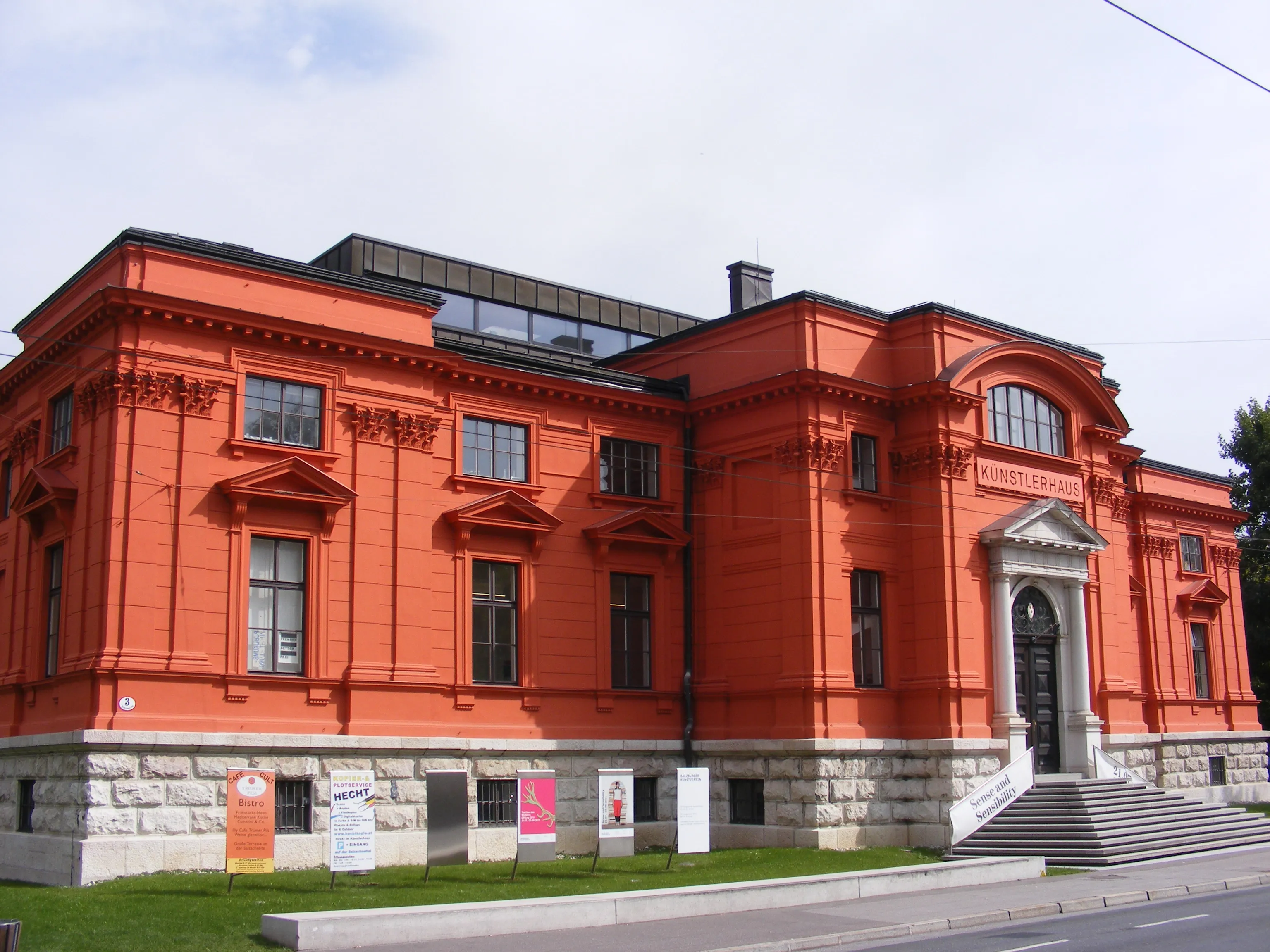 Histórico edificio Künstlerhaus de color rojo anaranjado con base de piedra, detalles de fachada clásicos y escalones de entrada, con carteles de exposiciones a lo largo de la planta baja