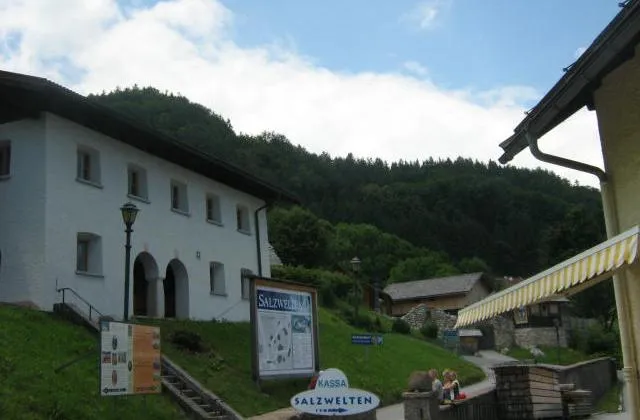 Vista exterior del centro de visitantes de la mina de sal Salzwelten con un edificio blanco, entradas arqueadas y una ladera boscosa al fondo.