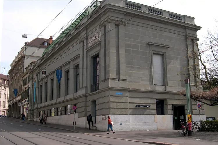 Hoekaanzicht van een groot neoklassiek stenen gebouw met blauwe spandoeken, tramrails op de voorgrond en voetgangers op het trottoir.