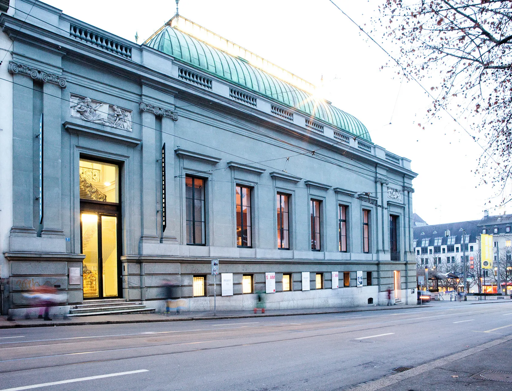 Stenen gevel van het Swiss Architecture Museum met een groen koperen koepeldak, grote ramen en een verlichte ingang langs een stadsstraat