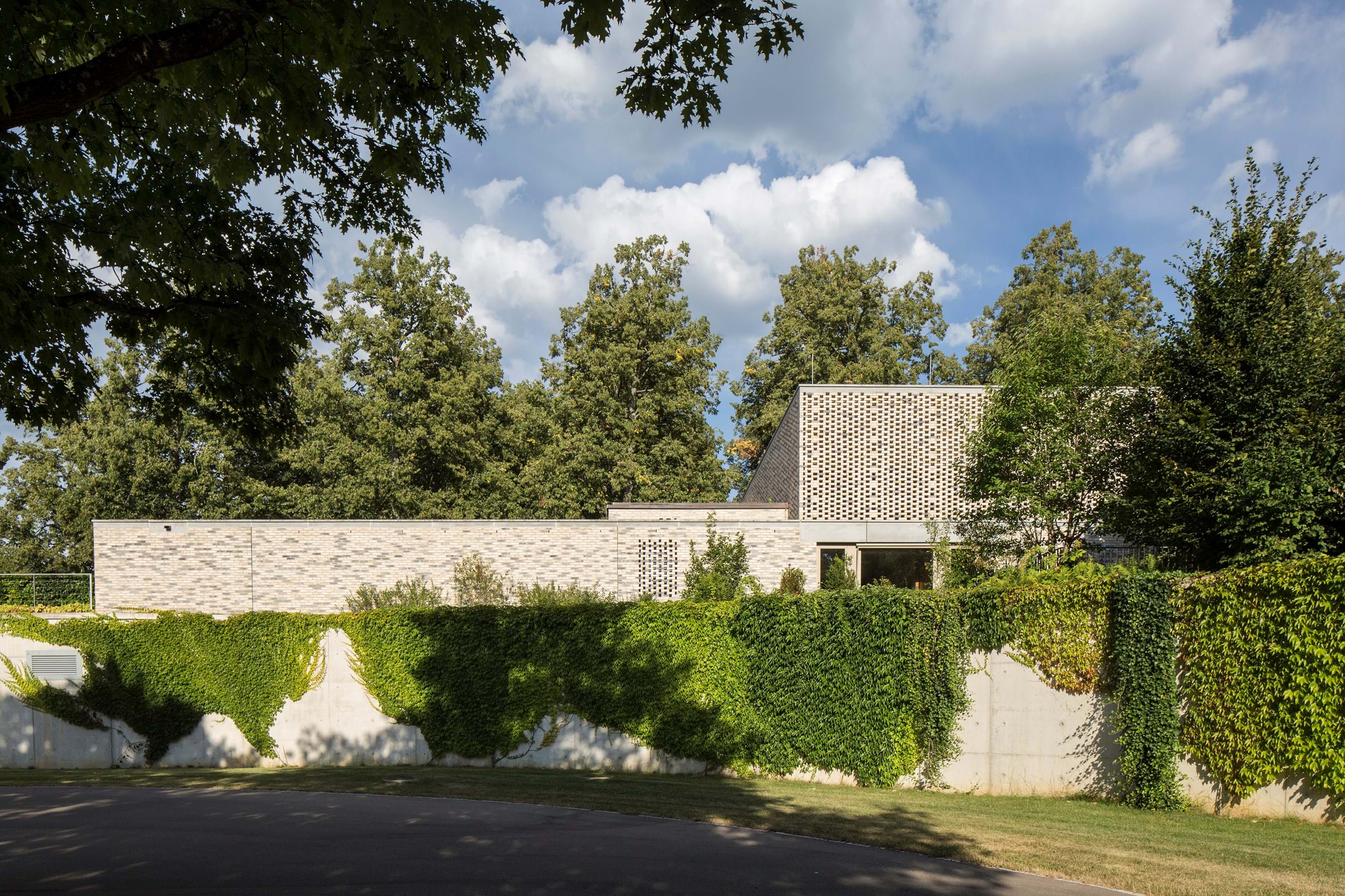 Een laag stenen gebouw met een getextureerde bakstenen toren die gedeeltelijk is bedekt met klimop, omringd door groene heggen en volwassen bomen onder een halfbewolkte hemel