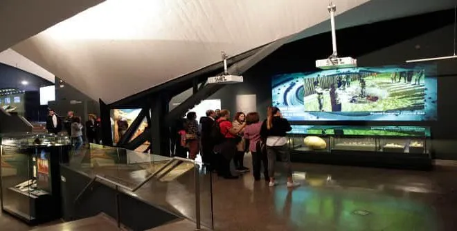 Grupo de personas viendo una pantalla grande y vitrinas en el interior de un museo