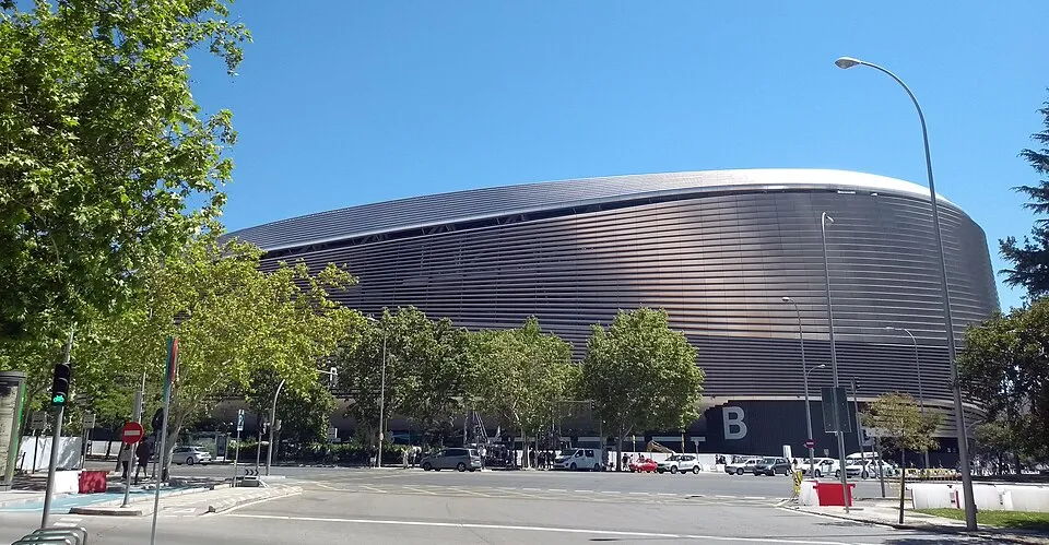 The Santiago Bernabéu Stadium with its curved metallic facade under a clear blue sky, surrounded by trees and a street.