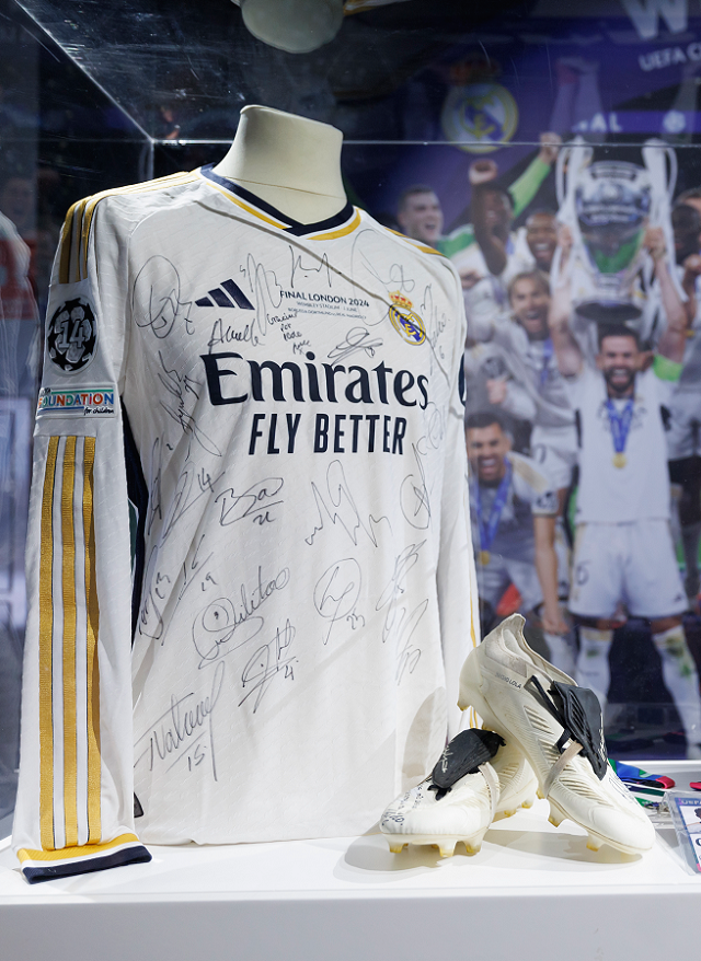 Signed white Real Madrid jersey with Emirates logo and signatures, displayed with football boots in a museum case
