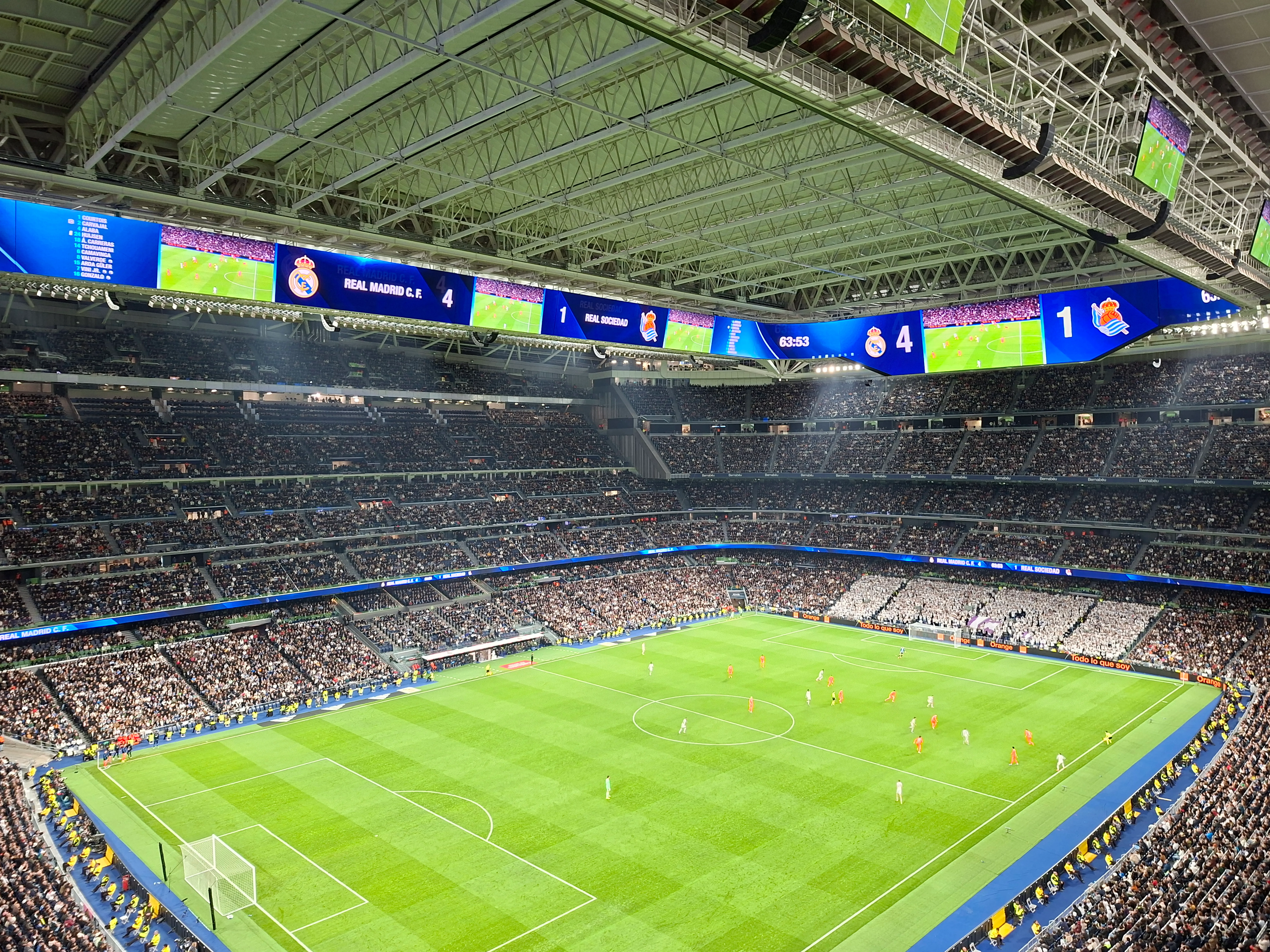 Wnętrze stadionu Santiago Bernabéu podczas meczu piłki nożnej, widok zielonej murawy, wypełnionych trybun i dużych cyfrowych tablic wyników