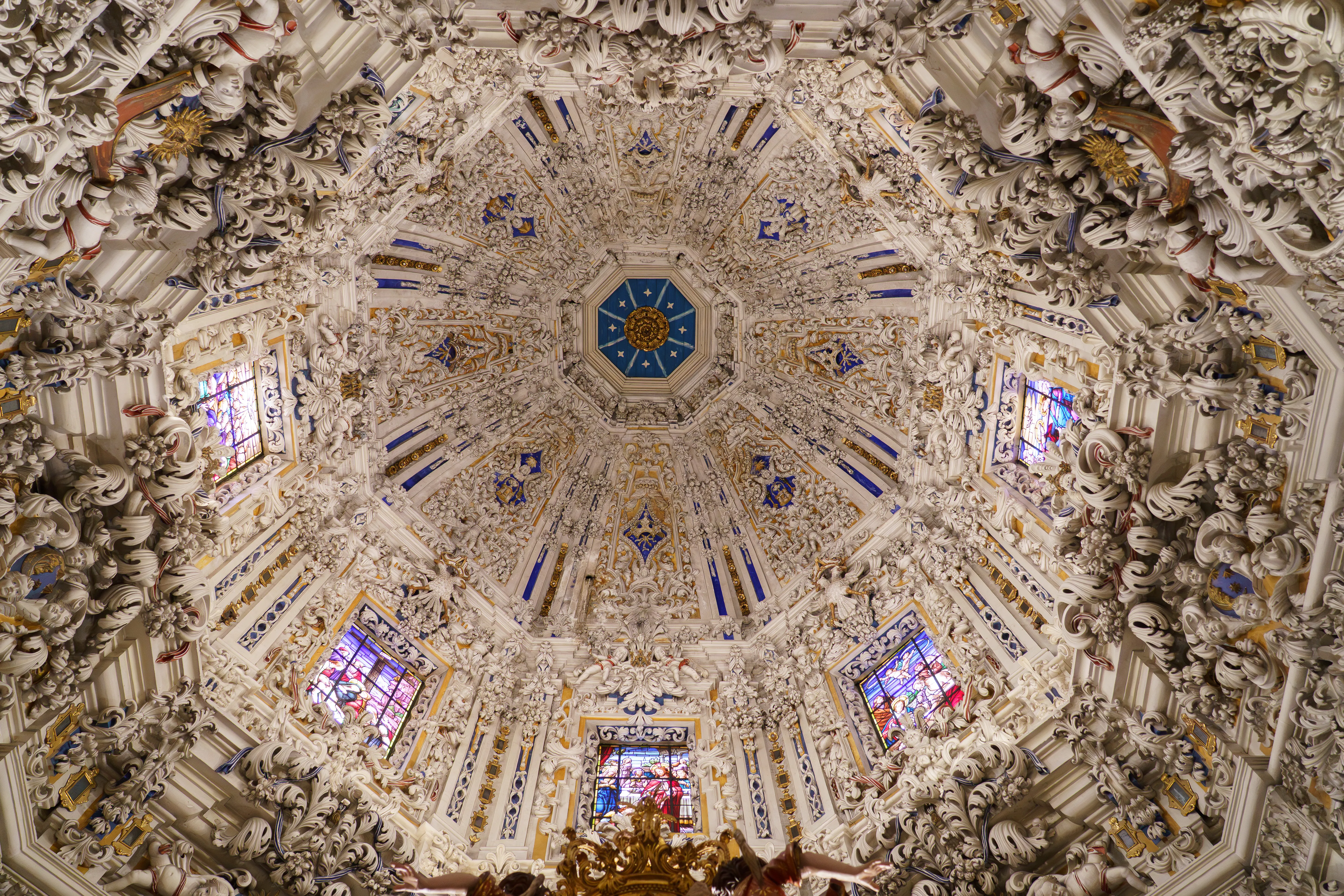 Rijkversierde barokke koepel met ingewikkelde witte stucwerkdecoraties, blauwe en gouden accenten en glas-in-loodramen in het Camarín van de Basilica of Santa María de la Victoria