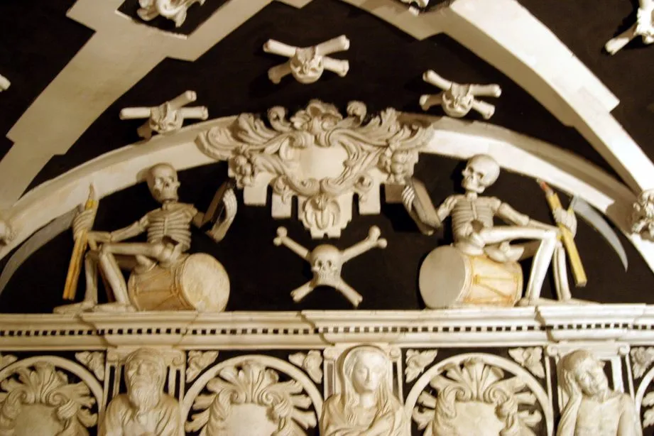 Wit barok stucwerk-reliëf met skeletten die trommelen, omringd door schedels en gekruiste beenderen op een zwarte achtergrond.