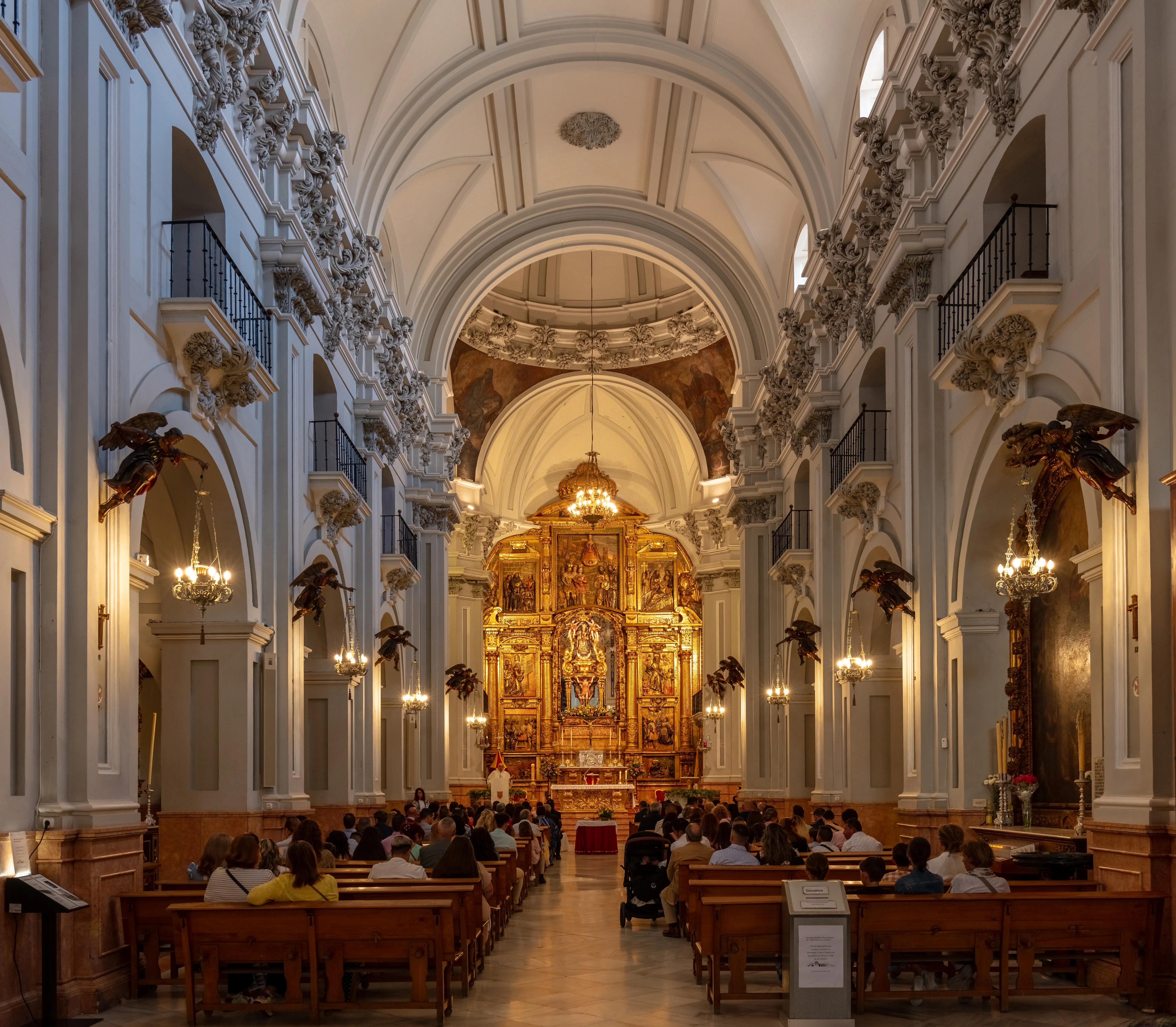 Interieur van een barokke basiliek met een gouden altaarstuk aan het einde, witte gewelfde plafonds, uitbundige wanddecoraties, kroonluchters en bezoekers die in houten kerkbanken zitten.