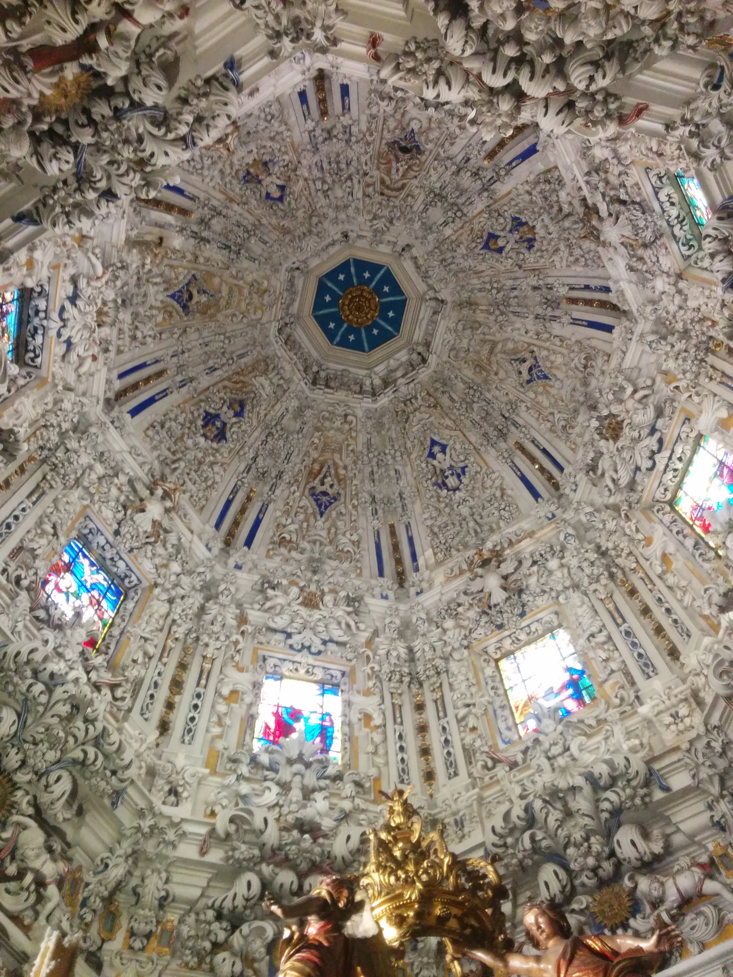 Ornate Baroque dome ceiling with intricate white stucco decorations, blue octagonal center medallion, stained glass windows, and golden sculptures at the base