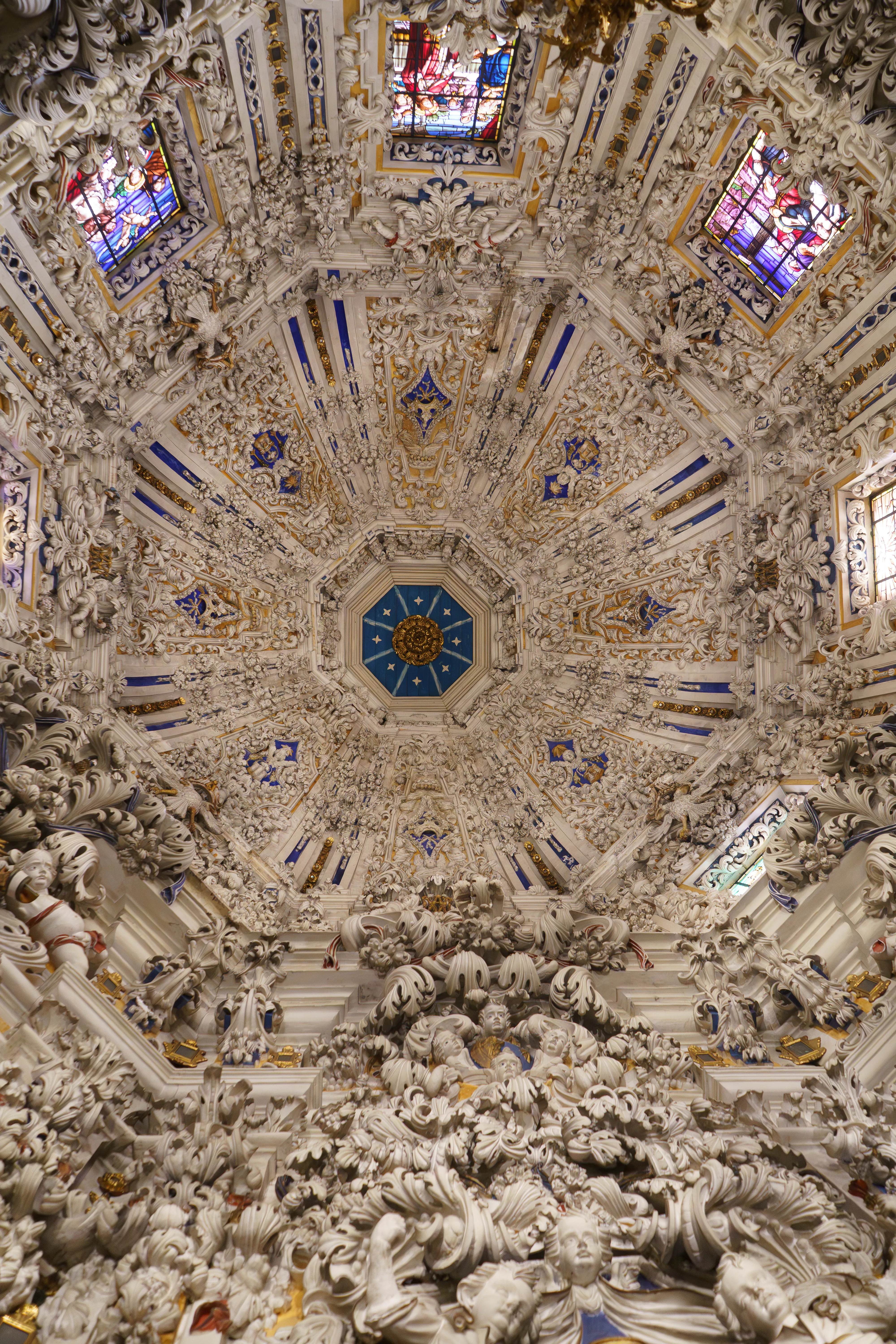 Omhoogkijkend naar de rijk versierde witte stucwerkkoepel met blauwe geometrische patronen en glas-in-loodramen.