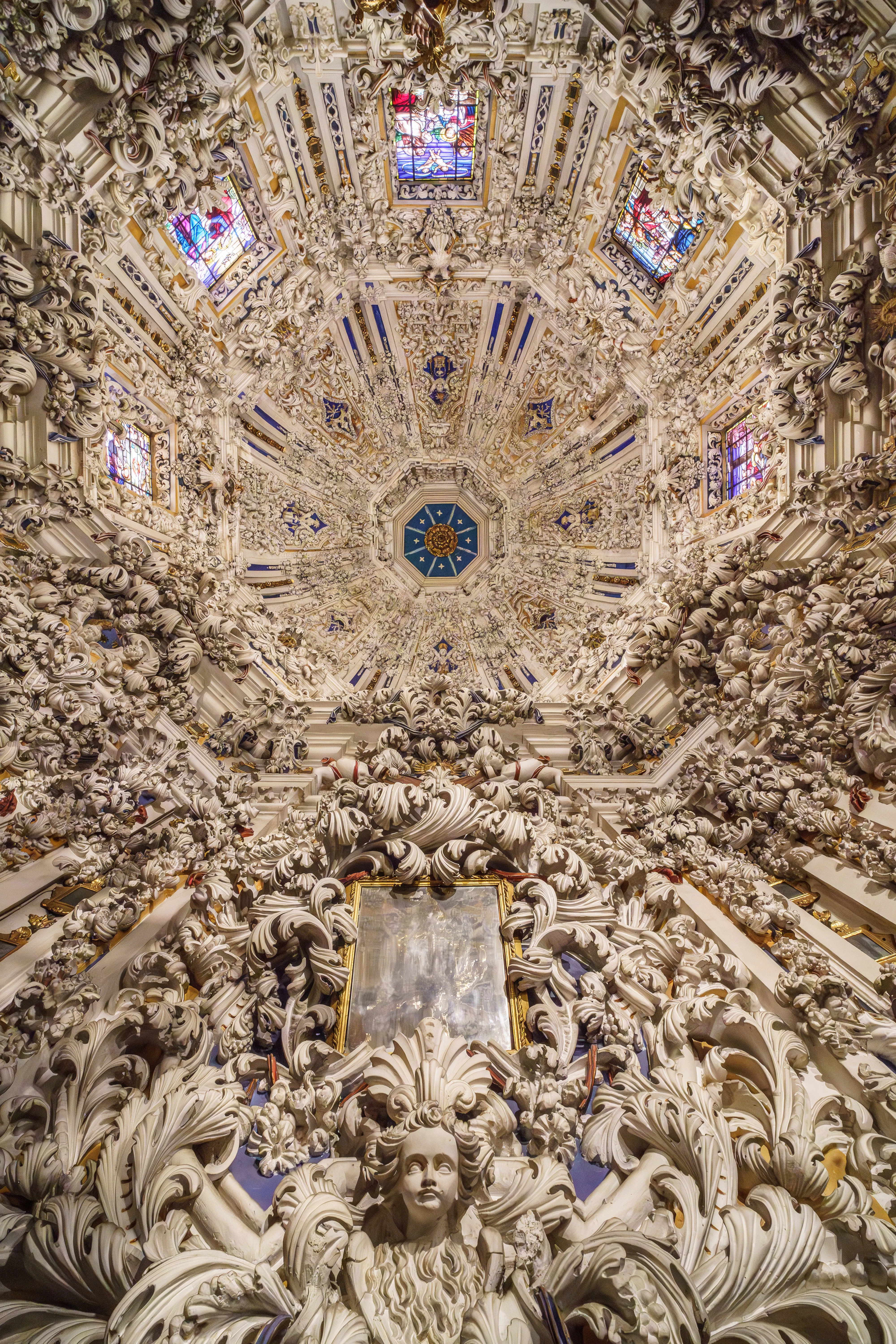 Omhoogkijkend naar het weelderige witte barokke stucplafond en de koepel van de Camarín, met gedetailleerde sculpturen, cherubijntjes en glas-in-loodramen.