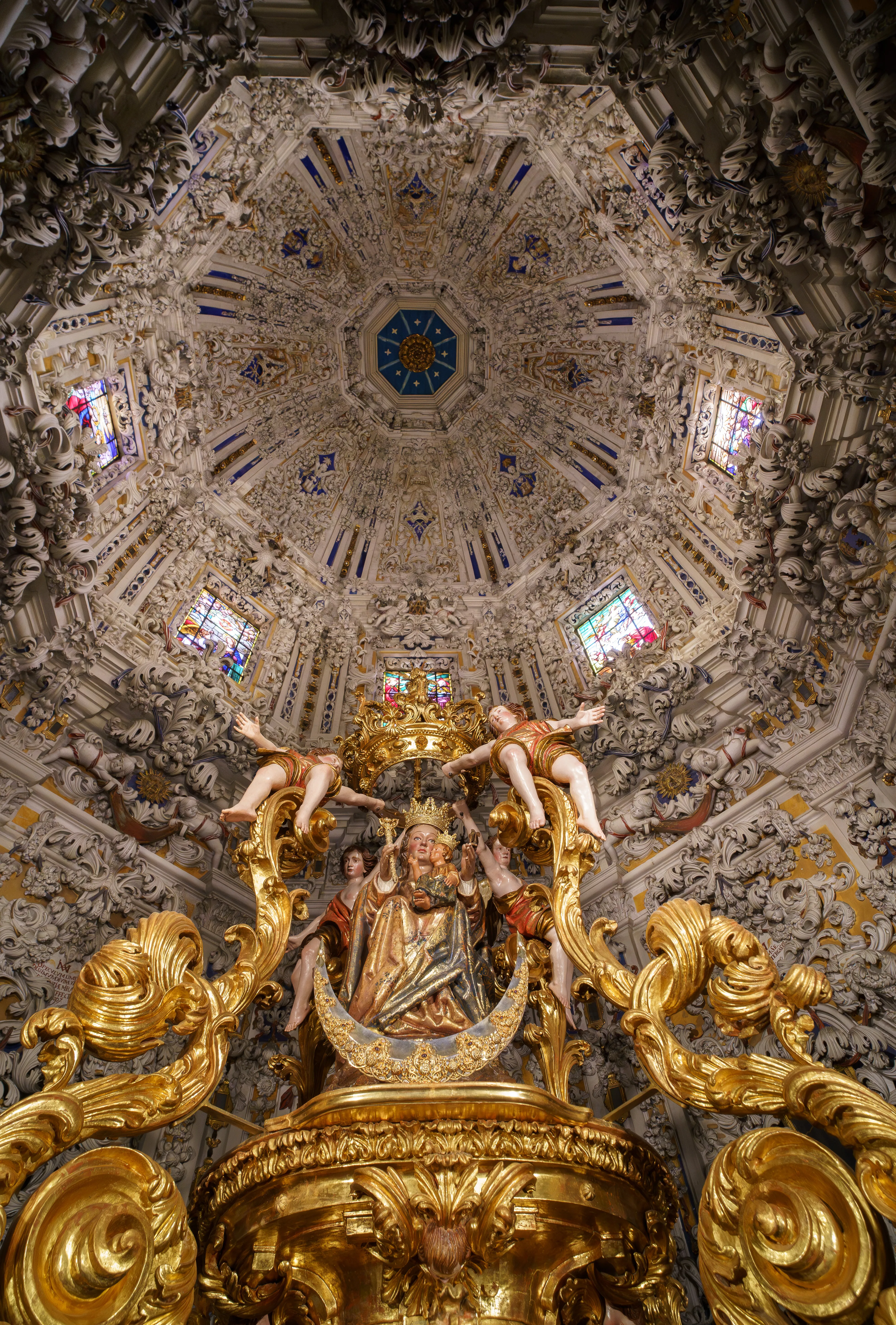 Kijk omhoog naar een rijkversierd barok koepelplafond met gedetailleerd wit stucwerk en een verguld altaarstuk met een beeld van de Maagd Maria en het kind, omringd door engelenfiguren.
