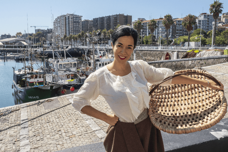 A smiling woman in a white blouse and brown skirt holds a large round wicker basket on a stone harbor quay, with fishing boats and waterfront buildings of Santurtzi port behind her