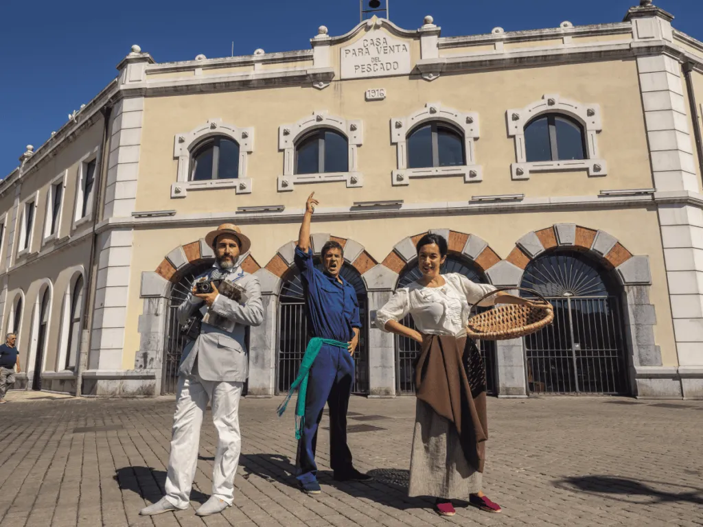 Three people in period costumes standing in front of the beige Museo Marítimo Pesquero building facade with arched windows and a blue sky
