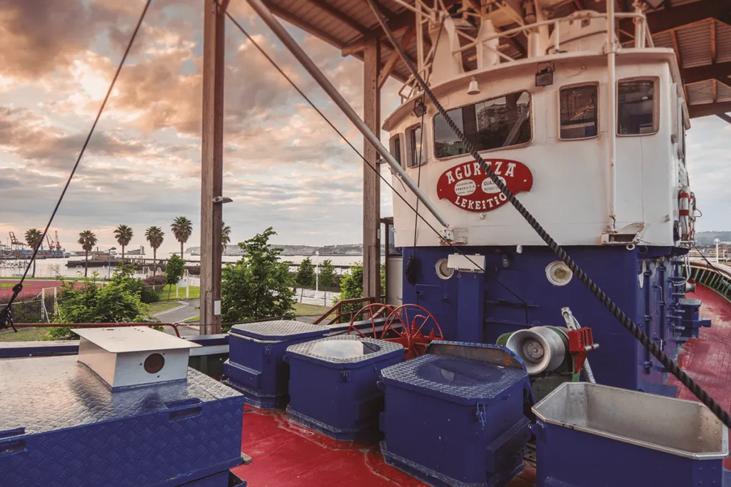 Deck of the Agurtza fishing boat with blue storage containers and red flooring under a shelter, with Santurtzi port and palm trees visible in the background