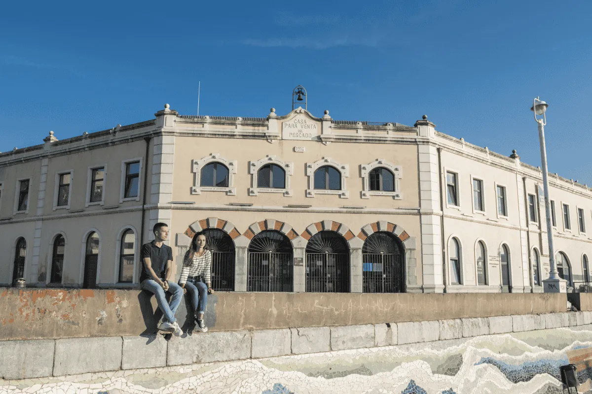 Two people sitting on a low wall in front of a large light-colored historic building with arched windows and iron-gated doorways under a clear blue sky