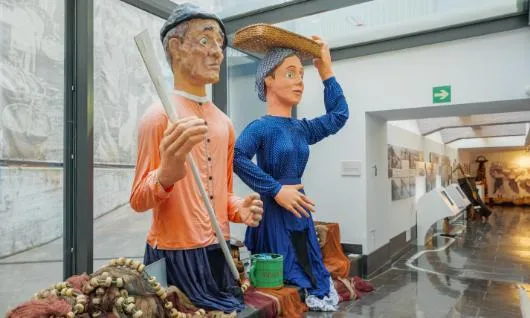 Two large painted figures of a fisherman in an orange shirt holding a pole and a woman in a blue dress carrying a basket on her head, displayed in a museum exhibition hall