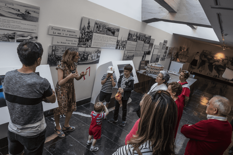 A group of adults and children on a guided tour inside a museum, viewing black-and-white historical photographs and text panels mounted on white walls beneath a skylight