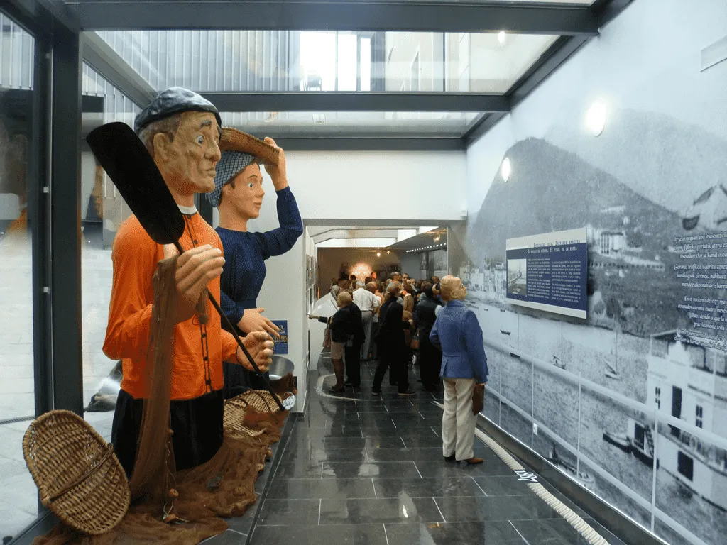 Two life-sized mannequin figures in traditional maritime clothing stand in a museum corridor, one holding a large oar, with visitors viewing a black-and-white harbor photograph on the wall behind them.