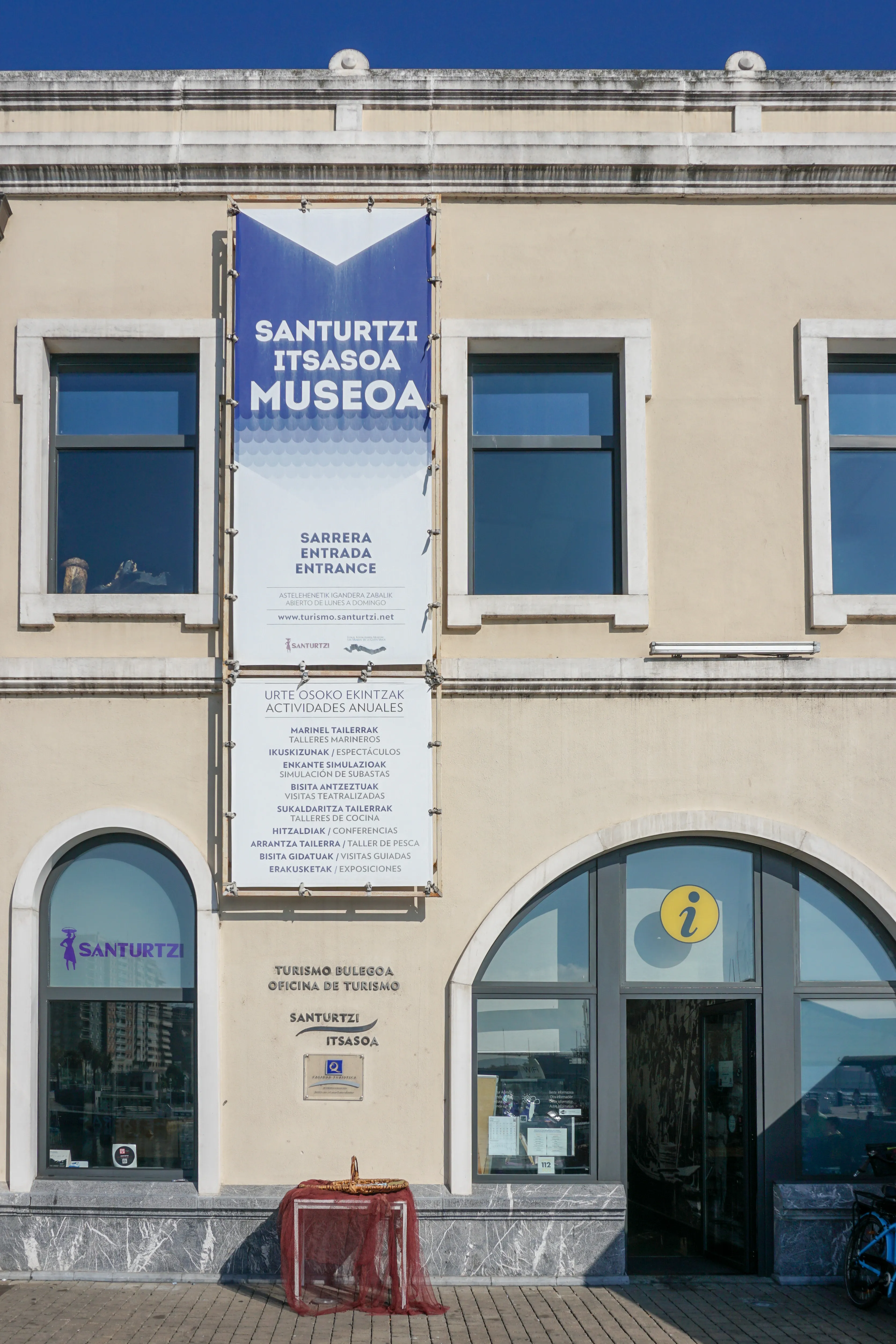 Beige building facade with a large vertical blue and white banner reading 'SANTURTZI ITSASOA MUSEOA' and 'ENTRANCE', arched ground-floor windows, and a tourist office sign