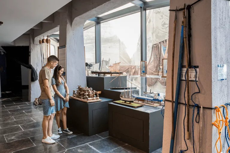 Two visitors stand inside the Santurtzi Museum of the Sea looking at model ship displays on dark pedestals, with maritime ropes and rigging hanging on the walls and large windows in the background.