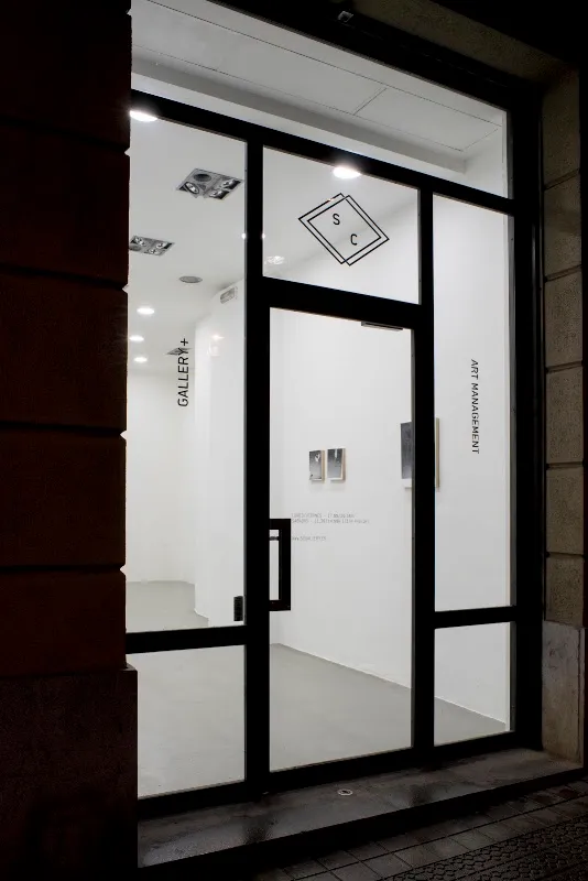 Puerta de entrada de cristal de SC Gallery con marco de metal negro, que muestra un logotipo de SC en forma de diamante sobre la puerta y el texto GALLERY+ y ART MANAGEMENT en los paneles de cristal.