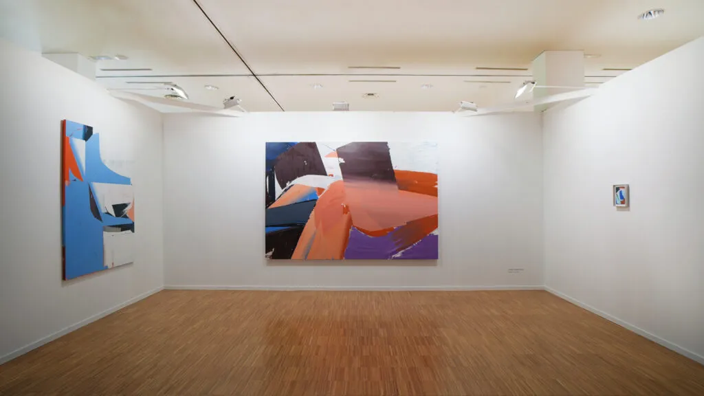 Una galería luminosa con paredes blancas y suelos de parqué de madera que exhibe grandes pinturas abstractas coloridas en tonos azules, naranjas y morados.