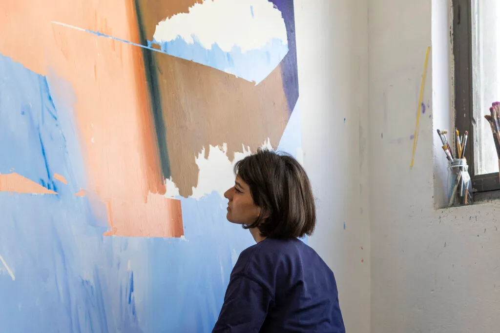 Una mujer de pelo corto y oscuro con una camisa azul marino mira hacia arriba un gran cuadro abstracto con bloques de color azul, melocotón y blanco en la pared de un estudio, con pinceles en un tarro en el alféizar de una ventana cercana
