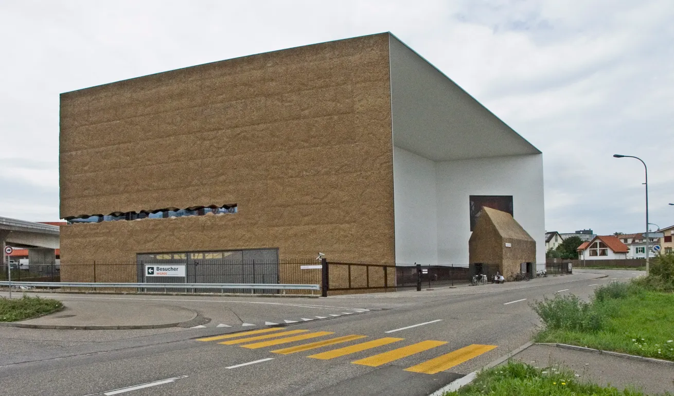 Widok z zewnątrz budynku Schaulager prezentujący charakterystyczną kanciastą fasadę z cegły i bieli, z ulicą i żółtymi oznaczeniami przejścia dla pieszych na pierwszym planie.