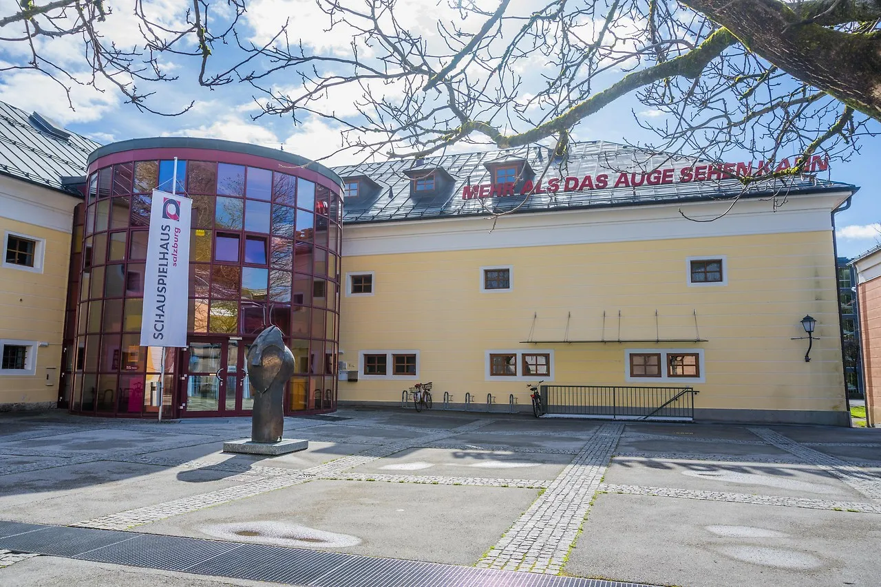 Widok z zewnątrz na Schauspielhaus Salzburg ukazujący nowoczesne szklane cylindryczne wejście dołączone do żółtego budynku, z rzeźbą z brązu na pierwszym planie i nagimi gałęziami drzew powyżej.