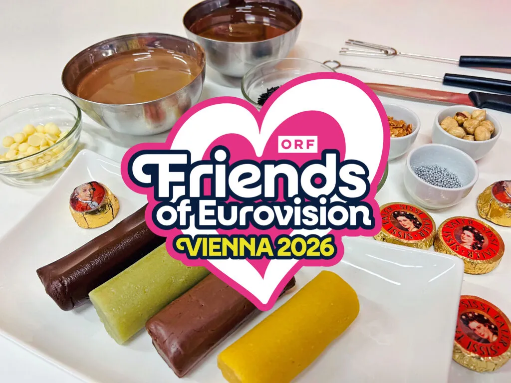 Tabliczki czekolady, miski z czekoladą i narzędzia do produkcji czekolady ułożone na białej powierzchni z nałożonym logo 'Friends of Eurovision Vienna 2026'