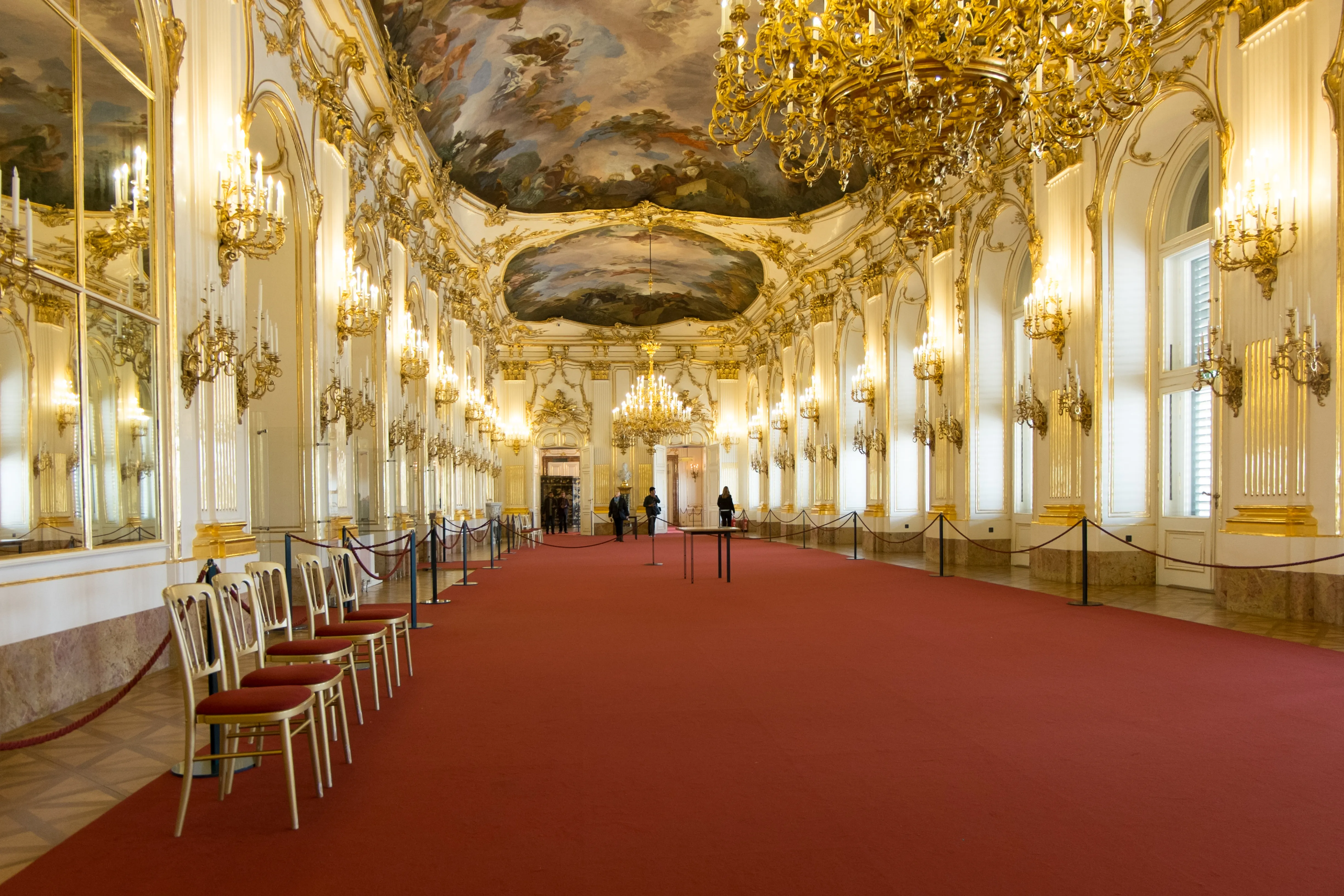 Ornate interior hall with red carpet, gold chandeliers, and a painted ceiling at Schönbrunn Palace