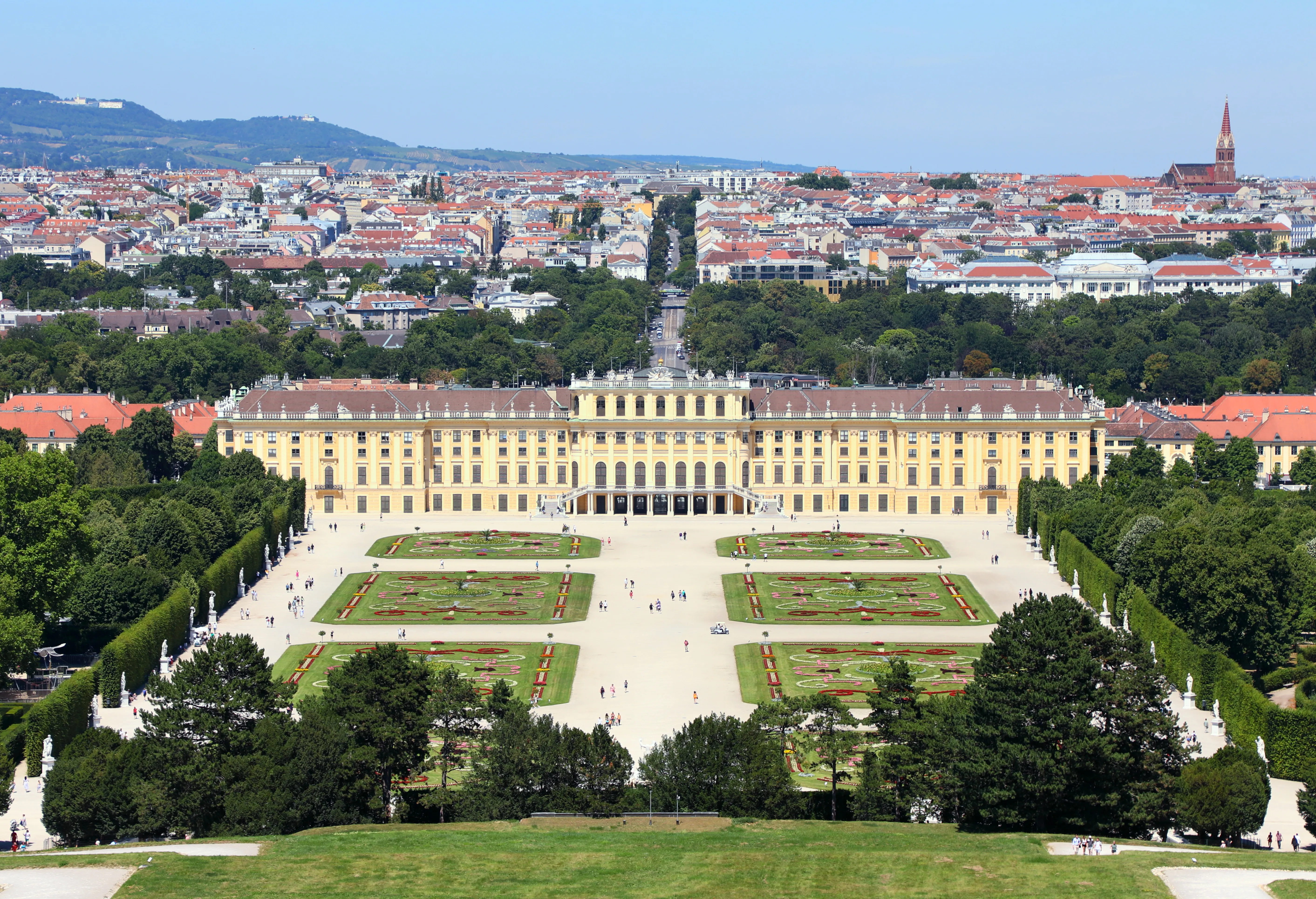 Elevated wide view of the yellow Schönbrunn Palace with formal parterre gardens in the foreground and the city of Vienna stretching into the distance.