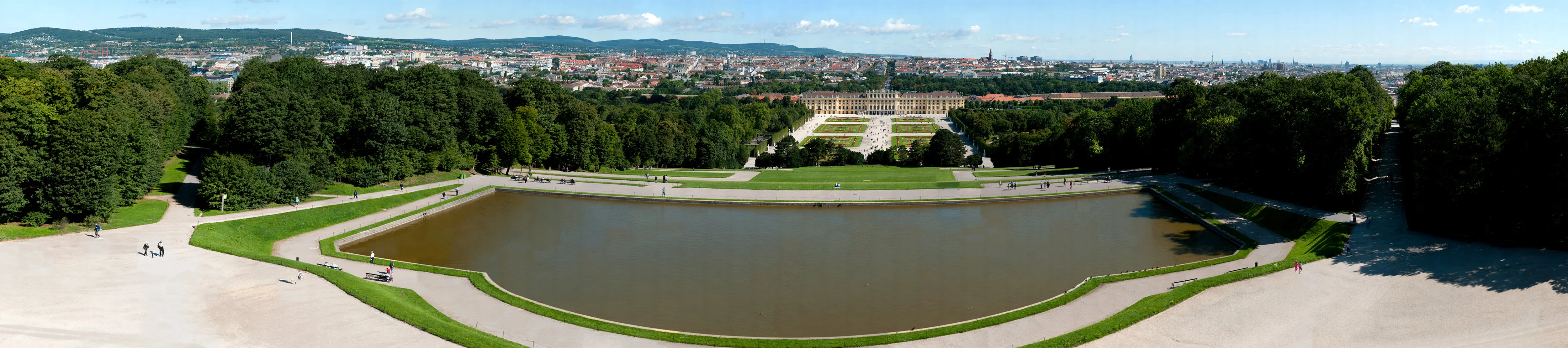 Panoramic view of Schönbrunn Palace with a large pond and green gardens in the foreground and the palace building in the distance.