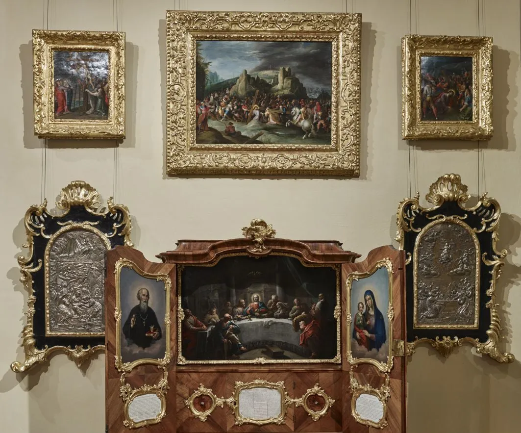 ornate schilderijen in vergulde lijsten en een houten altaarstuk aan een museummuur
