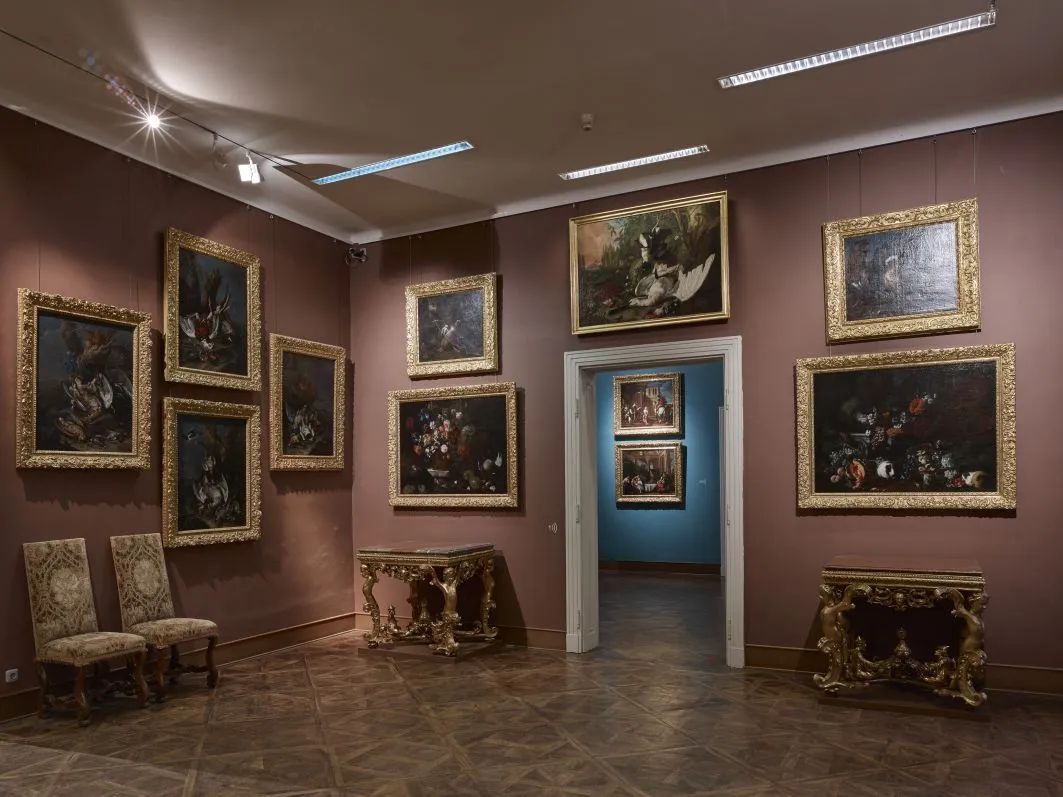 Galerijkamer met schilderijen in vergulde lijsten aan miauwwitte muren, verguld meubilair en een deuropening naar een andere kamer