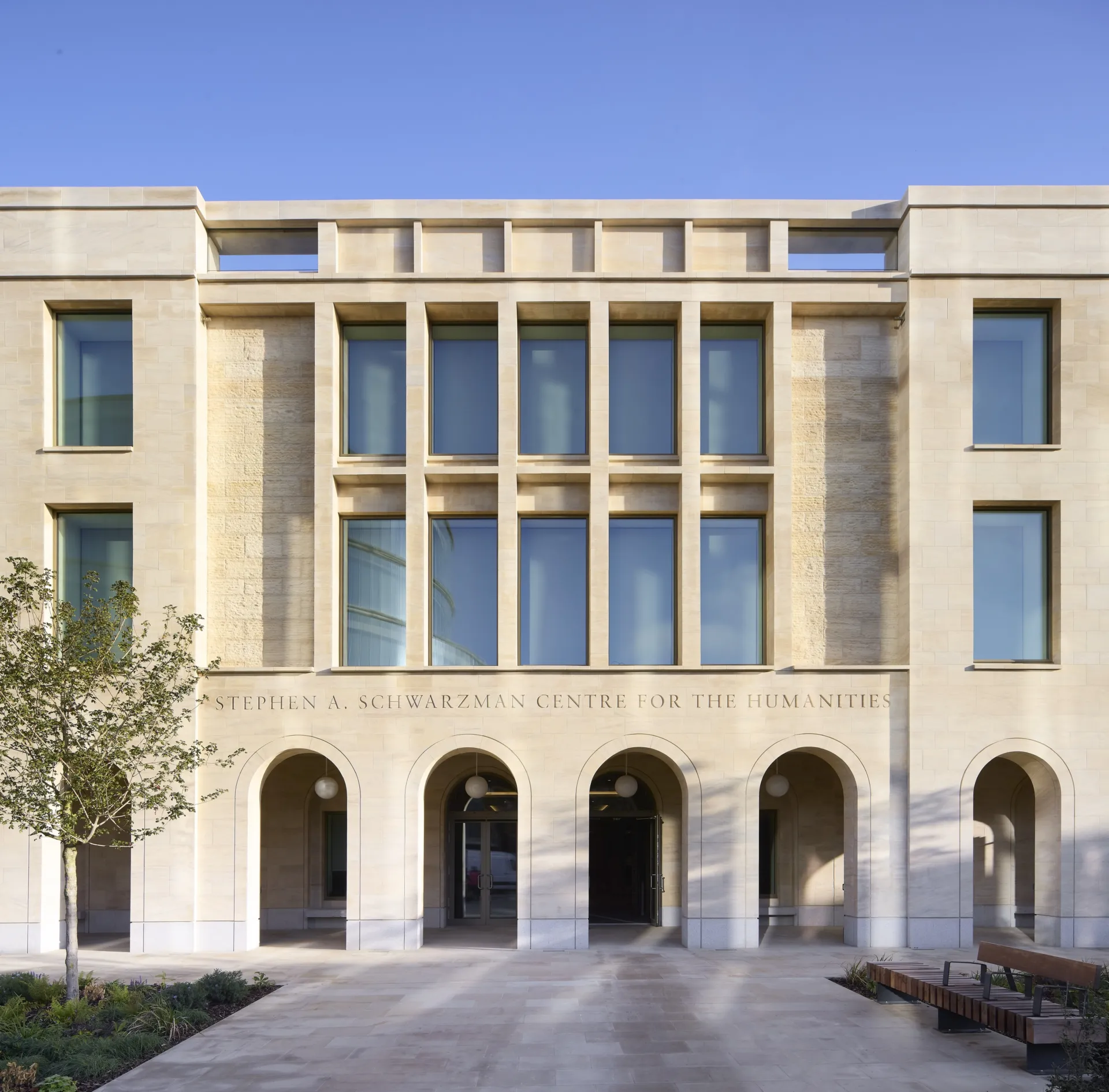 Stone facade of the Stephen A. Schwarzman Centre for the Humanities with arched entrance portico and large windows under a clear blue sky