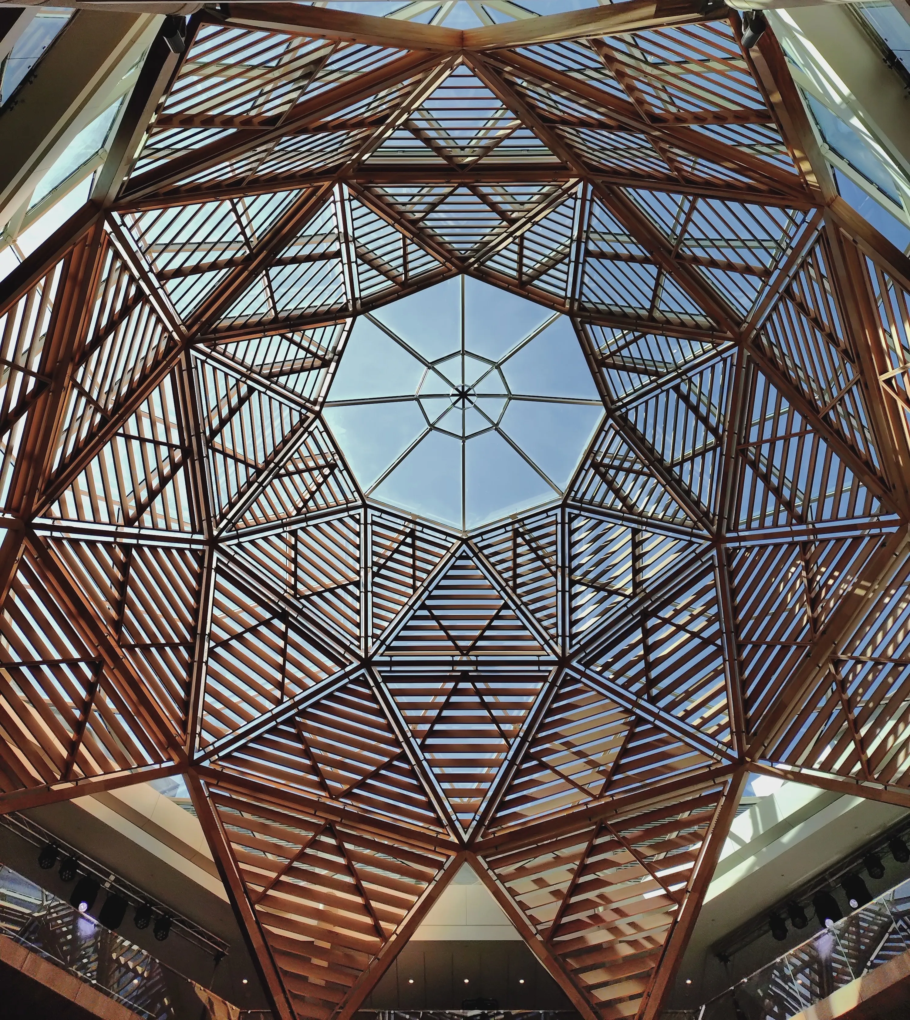 Looking up at a geometric timber lattice atrium ceiling with a hexagonal skylight at the center, showing intricate wooden beam framework against a blue sky.