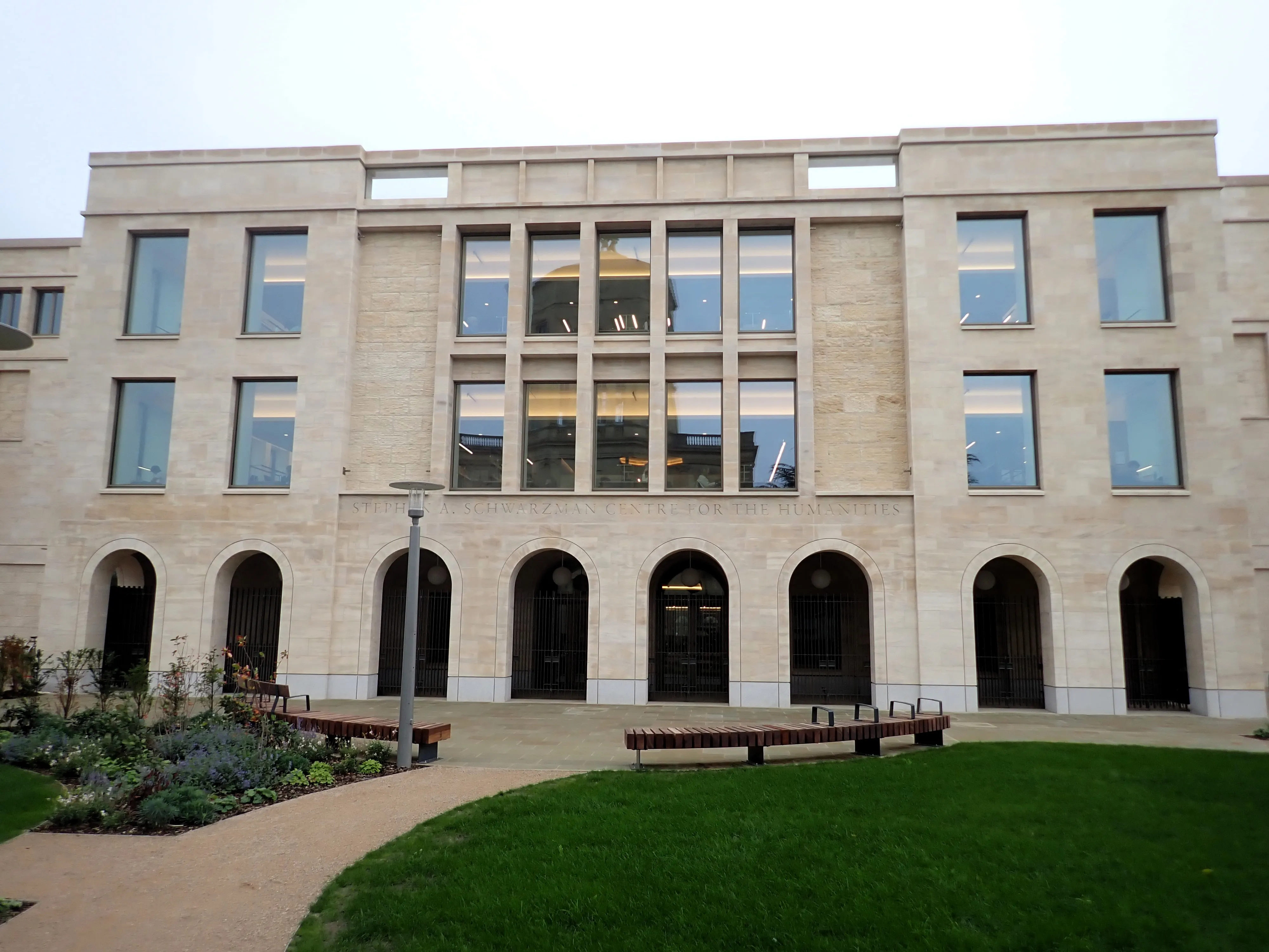 Stone facade of the Stephen A. Schwarzman Centre for the Humanities with arched ground-floor entrances, large upper windows, and a curved lawn with benches in front