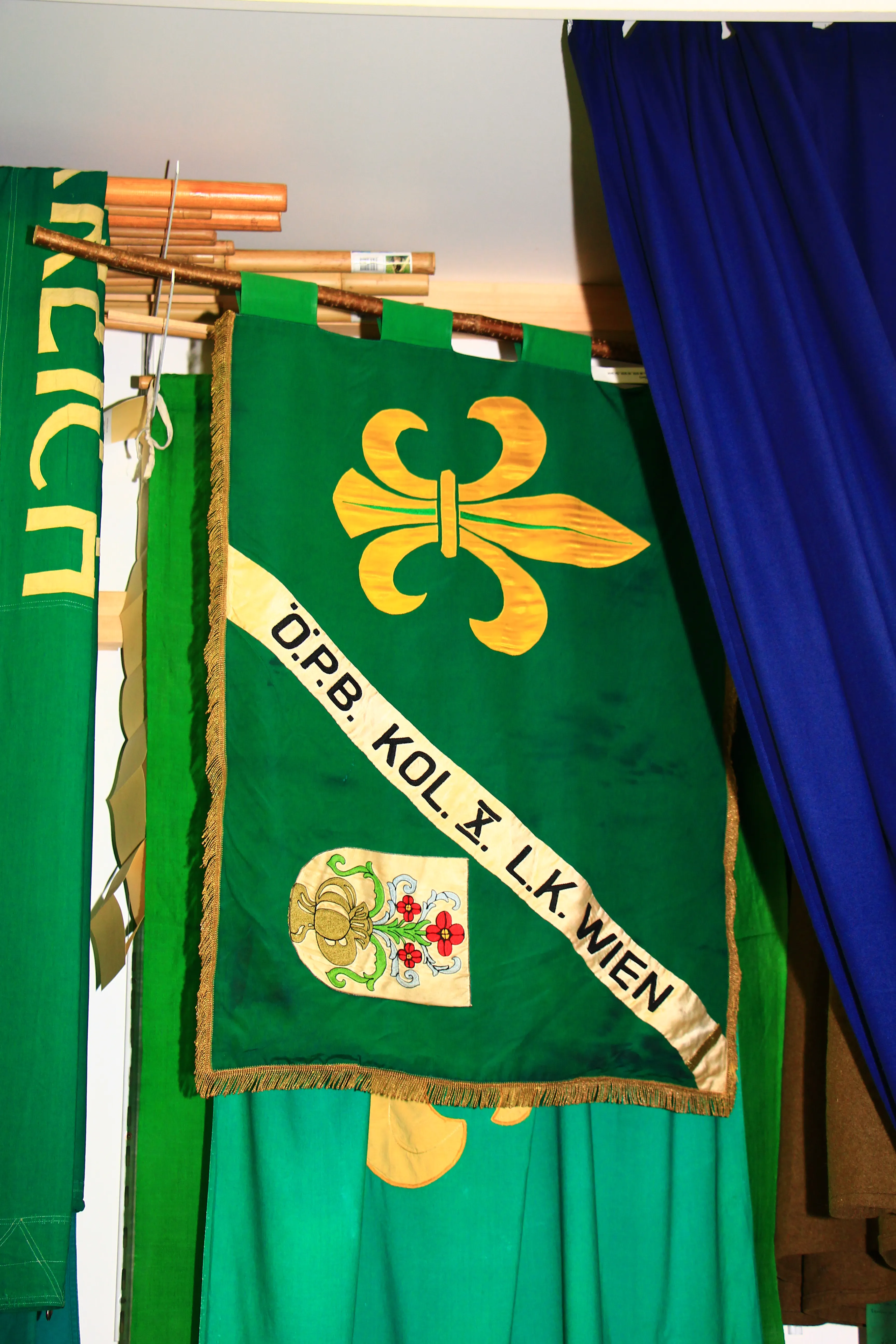 Green banner with yellow fleur-de-lis and text 'Ö.P.B. KOL. X. L.K. WIEN' displayed indoors