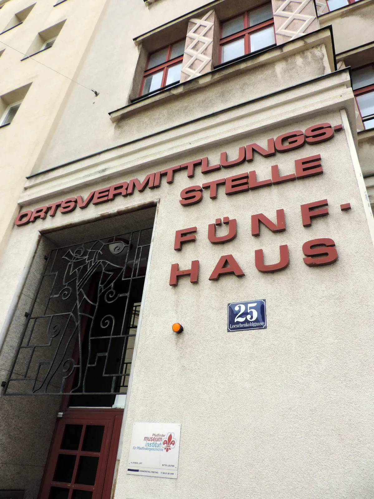 Fasada budynku z czerwonym znakiem z napisem ORTSVERMITTLUNGSSTELLE FÜNFHAUS i numerem 25