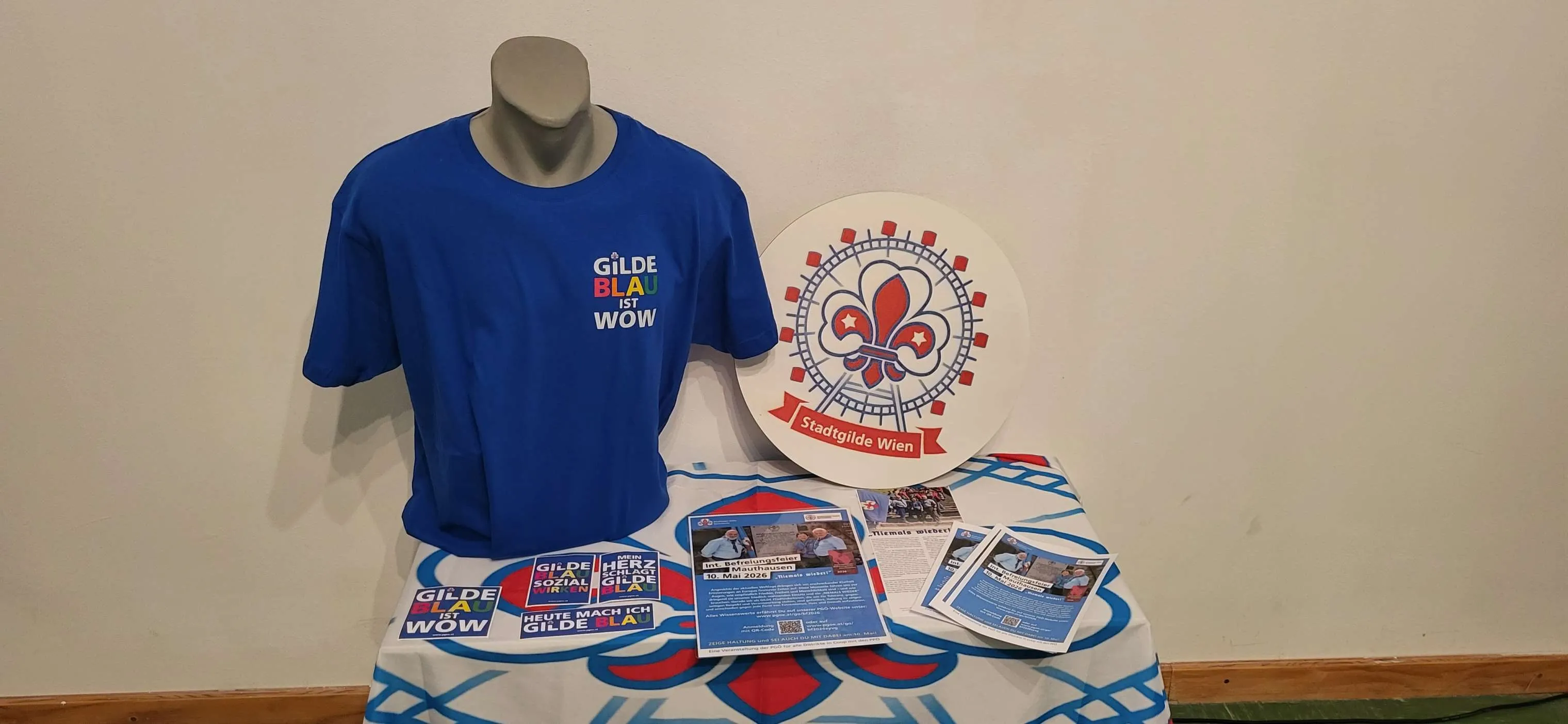 Blue t-shirt with 'GILX BIA WOW' text, a circular emblem with a fleur-de-lis, and brochures on a patterned tablecloth