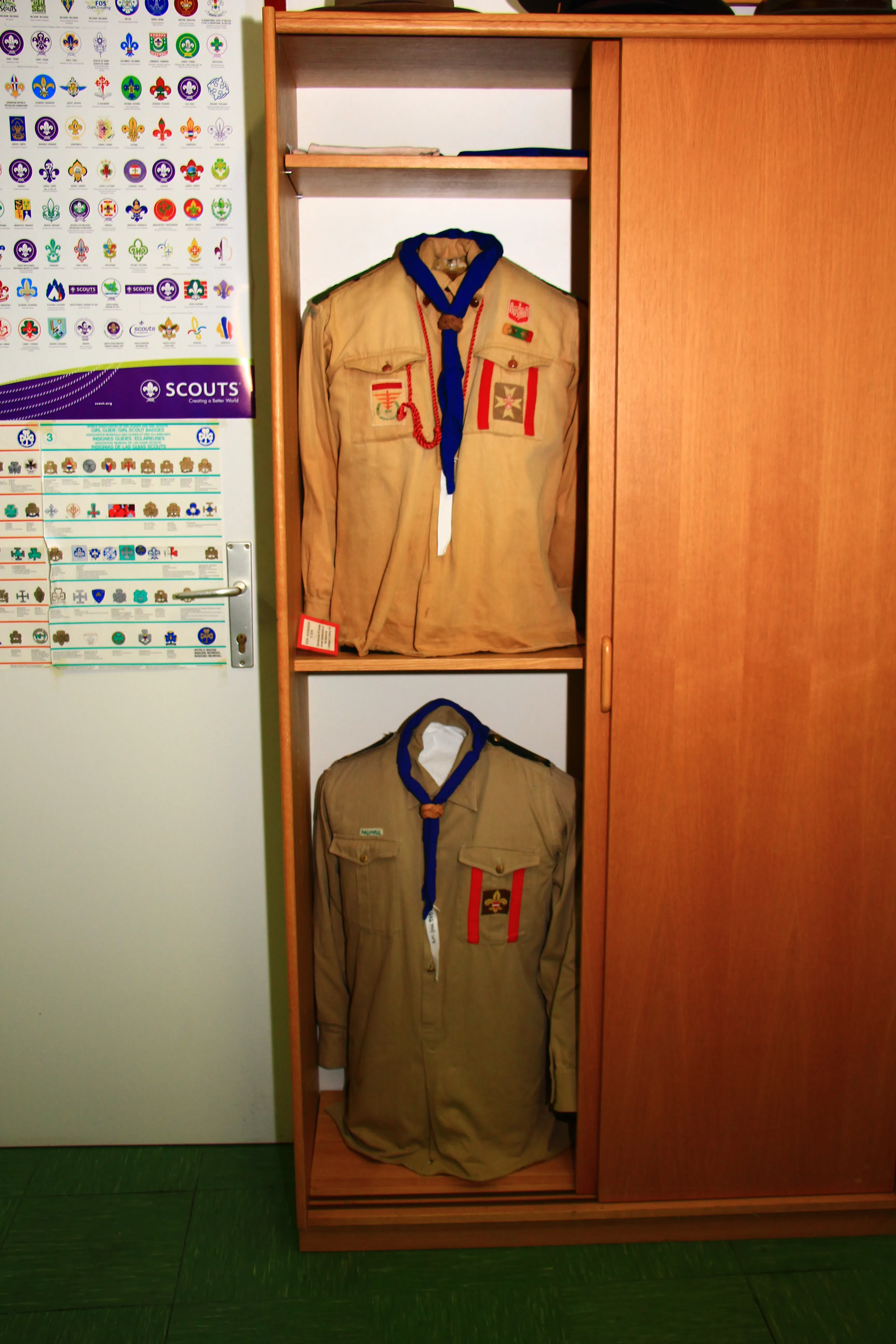 Two scout uniforms displayed in a wooden cabinet with a wall chart of scout badges