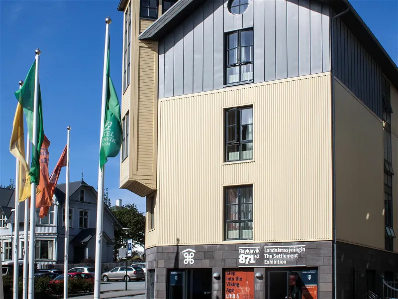 Widok z zewnątrz na budynek The Settlement Exhibition w Reykjavíku, ukazujący wielopiętrową fasadę z blachy falistej i drewnianą okładziną, duże okna oraz kilka kolorowych flag na masztach przed wejściem z widocznym oznakowaniem.