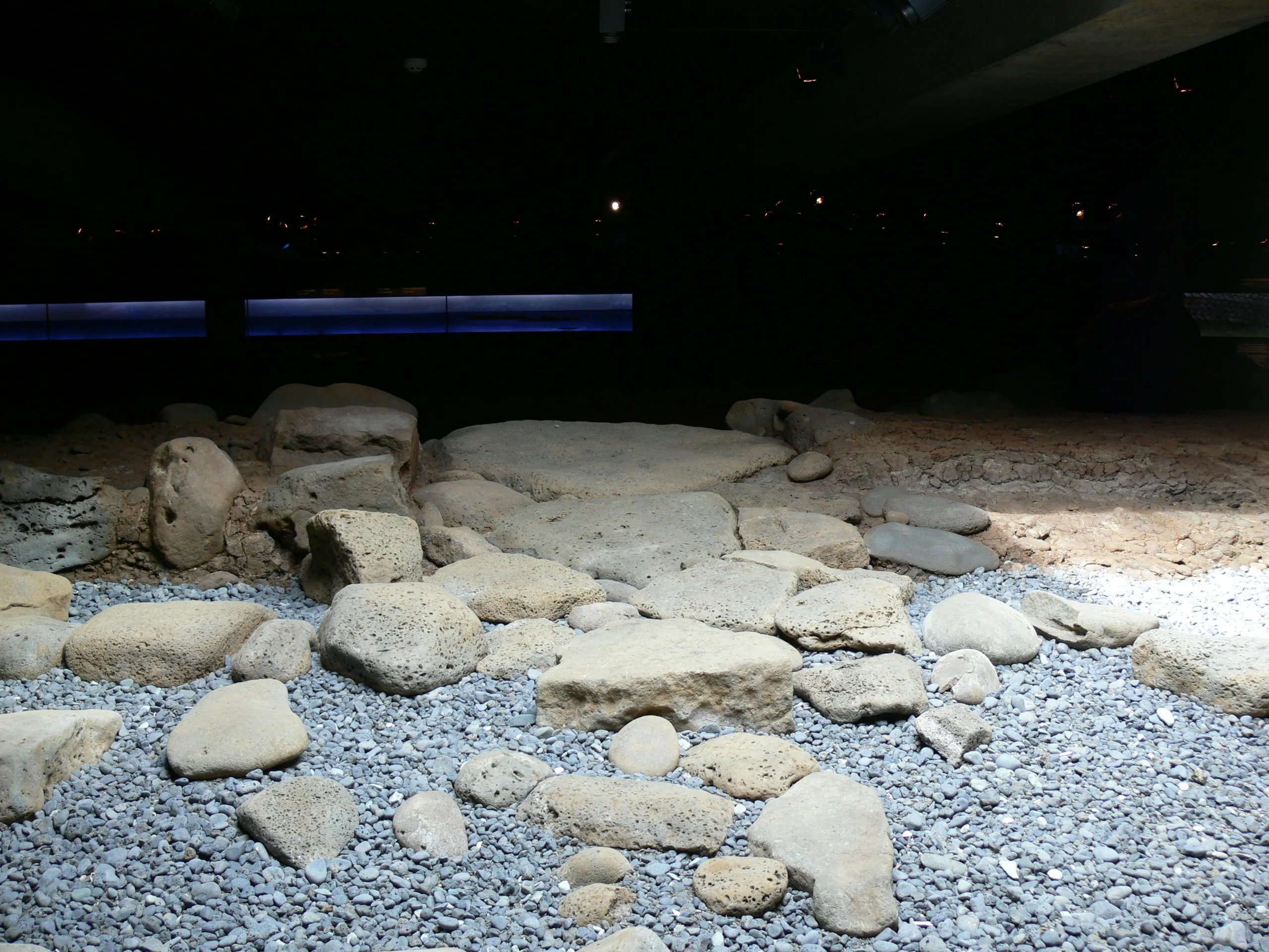 Wystawa wykopalisk archeologicznych z rozrzuconymi jasnymi kamieniami i skałami na szarym żwirze oraz niebieskimi paskami oświetlenia LED widocznymi na tle ciemnej ściany