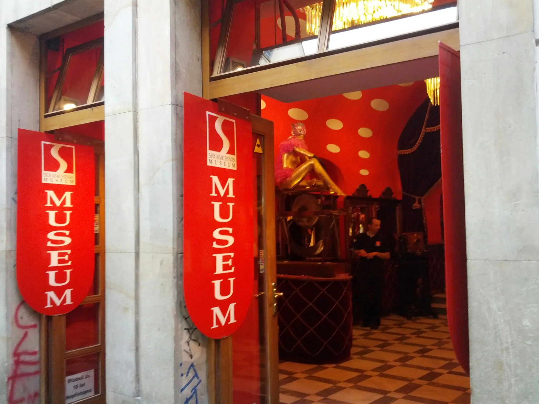 Rode spandoeken met de tekst 'MUSEUM' en een logo bij de ingang van een stenen gebouw, met uitzicht op een interieur met rode muren.