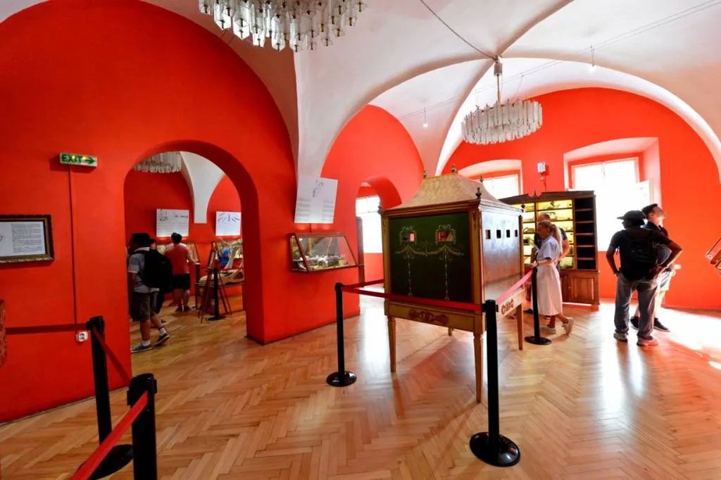 Interieur van een museum met rode muren, gewelfde plafonds en bezoekers die tentoonstellingen bekijken achter barrières