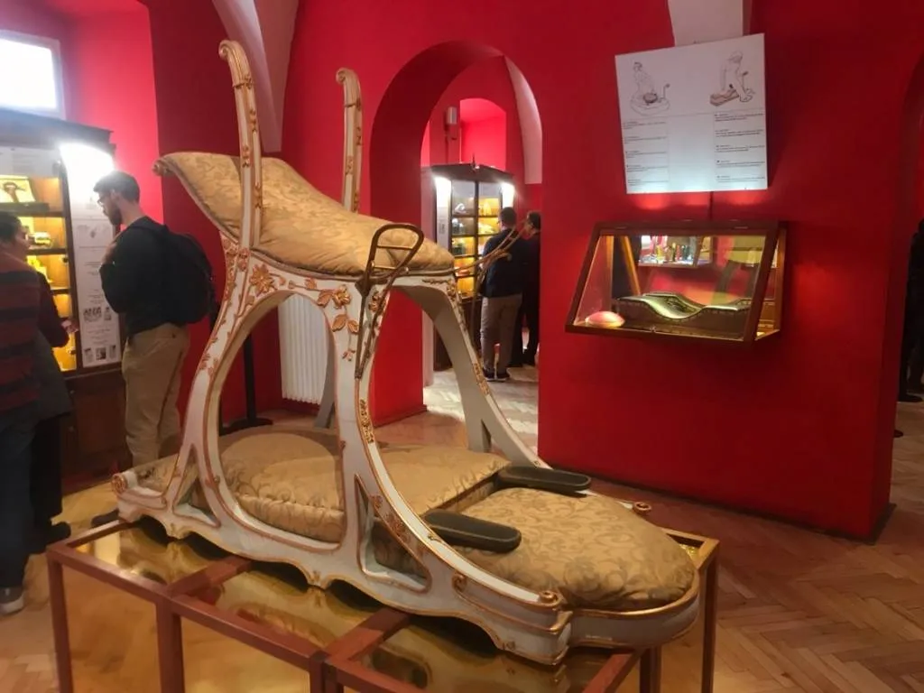 Sierlijke vintage seksmachine met gestoffeerde zitting en mechanische componenten tentoongesteld in een museumgalerij met rode muren.